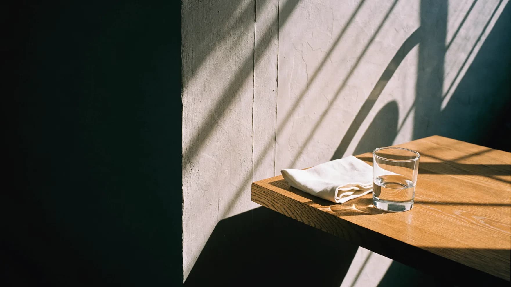 De hoek van een eiken tafel in hard ochtendlicht, met een gevouwen linnen servet en een glas water; lange schaduwen over een pleisterwand